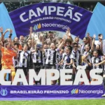 O time feminino do Corinthians celebra a vitória no Campeonato Brasileiro, com jogadoras levantando a taça no palco, em frente a uma grande faixa com a palavra "Campeãs".