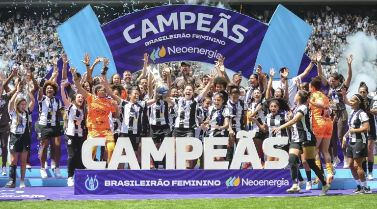 O time feminino do Corinthians celebra a vitória no Campeonato Brasileiro, com jogadoras levantando a taça no palco, em frente a uma grande faixa com a palavra "Campeãs".